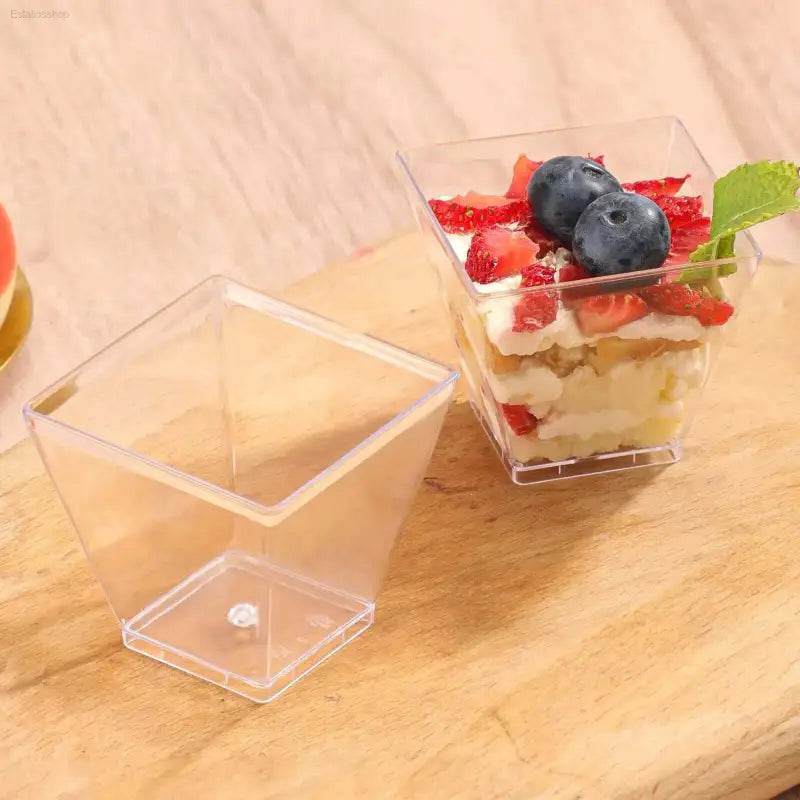 Two clear, square dessert cups, one filled with layered cake, cream, strawberries, blueberries, and a mint leaf.
